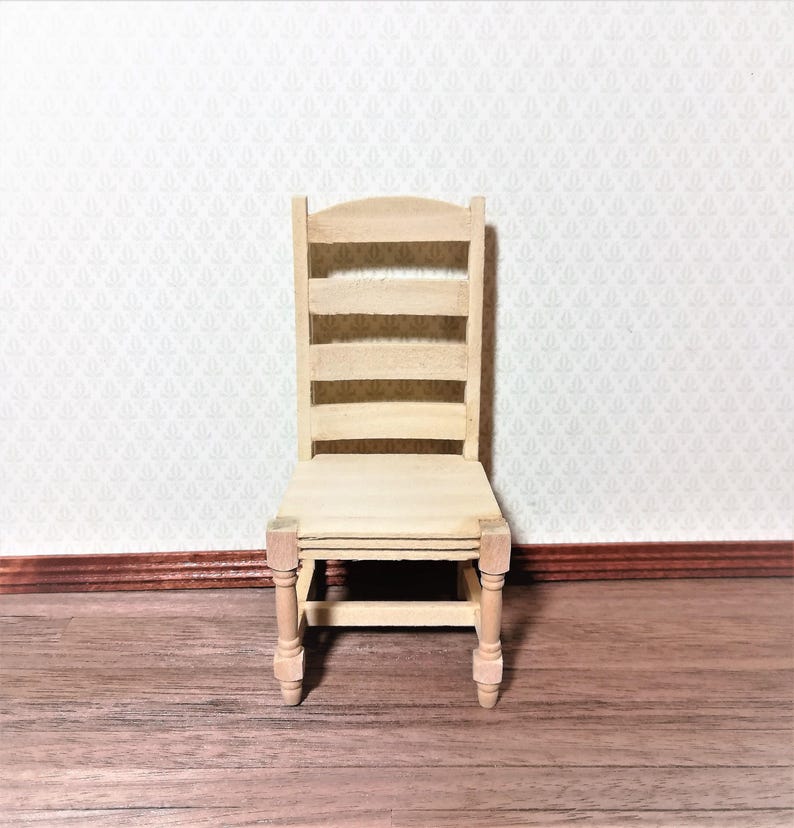 May include: A miniature wooden chair with a ladder back design. The chair is unfinished, showing the natural wood grain. The seat and legs are also made of wood. The chair is set against a patterned wallpaper background and a wooden floor.
