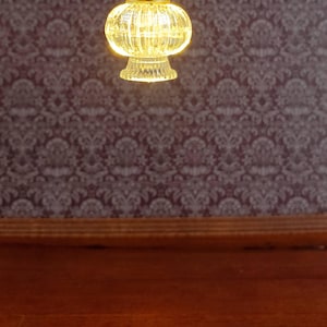 May include: A miniature, glowing lamp with a clear, ribbed glass shade and a gold-colored metal fixture. The lamp is suspended against a patterned, dark purple wallpaper and a wooden surface.