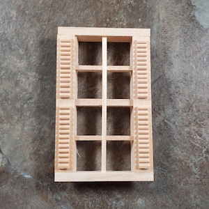 May include: A miniature wooden window with shutters. The window frame is rectangular with eight panes. The shutters are on either side of the window and have a slatted design. The wood is unfinished and has a light, natural color.