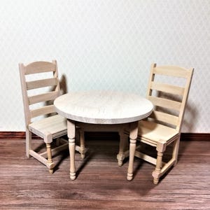 May include: A miniature wooden dining set comprising a round table and two chairs. The table, with a light wood finish, has four turned legs. The chairs feature a ladder-back design, also in a light wood tone. Suitable for dollhouses.