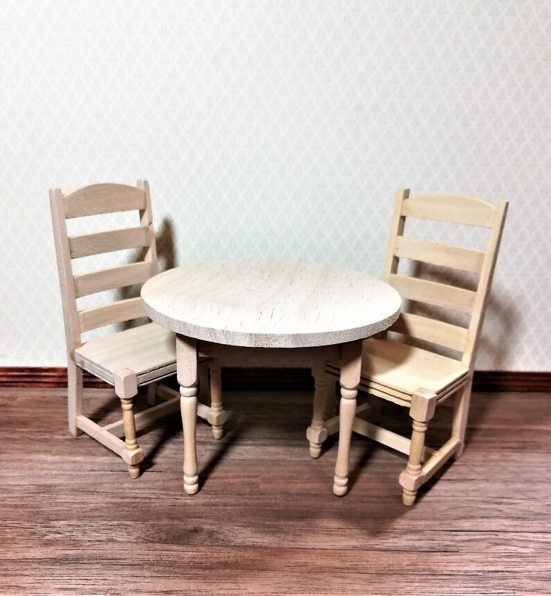 May include: A miniature wooden dining set comprising a round table and two chairs. The table, with a light wood finish, has four turned legs. The chairs feature a ladder-back design, also in a light wood tone. Suitable for dollhouses.