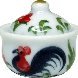 Könnte beinhalten: Eine kleine weiße Keramikschale mit Deckel, verziert mit einem handgemalten Hahn-Motiv in Schwarz und Rot sowie grünen Blattakzenten. Der Deckel hat einen abgerundeten Knauf und passende florale Muster auf einer abgerundeten Basis.