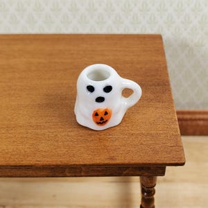 Könnte beinhalten: Ein Miniatur-Becher aus weißer Keramik in Form eines Geistes mit einem kleinen Henkel. Der Geist hat zwei schwarze, kreisförmige Augen, einen schwarzen, kreisförmigen Mund und hält einen kleinen orangefarbenen Kürbis mit einem schwarzen Gesicht.
