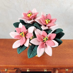 May include: A miniature poinsettia arrangement with pink and white petals, yellow centers, and dark green leaves. The flowers are in a small yellow pot, set on a wooden surface. The arrangement is a decorative item.