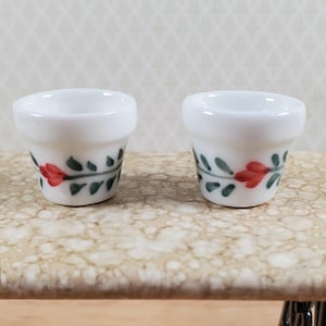May include: Two miniature white ceramic flower pots featuring a hand-painted floral motif with small red petals and green leaves encircling the exterior. Each pot has a wide rim and a tapered base, designed with a smooth, glossy finish.