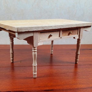 May include: A miniature wooden table with two small drawers and turned legs. The table is light-colored with a natural wood grain finish. The table is approximately 10 inches long and 8 inches wide.