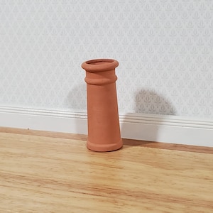 May include: A miniature terracotta chimney pot. The small, cylindrical pot has a flared top and a slightly tapered body. The chimney pot is a warm, reddish-brown color and is likely made of clay. It is designed for a dollhouse or miniature scene.