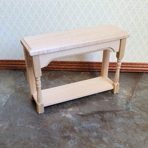 May include: A miniature, unfinished wooden console table. It features a rectangular top and a lower shelf, supported by decorative turned legs. The table is designed for dollhouses or similar settings. The natural wood finish provides a neutral aesthetic.