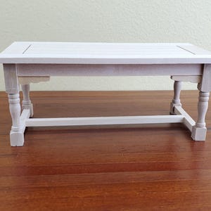 Op de afbeelding: Een kleine, witte houten tafel met een rechthoekig blad en vier gedraaide poten. De tafel is ongeveer 61 cm lang, 30 cm breed en 25 cm hoog. De tafel is een miniatuurmeubelstuk.