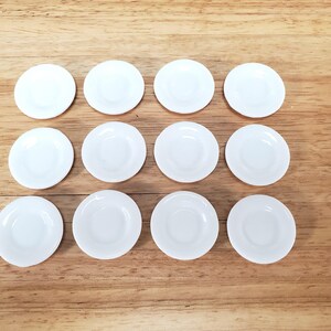 Pu&ograve; includere: Un set di dodici piccoli piattini circolari in ceramica bianca disposti in una griglia di tre file e quattro colonne. Ogni piattino presenta una finitura liscia e lucida con un centro leggermente incavato e un bordo rialzato uniforme.