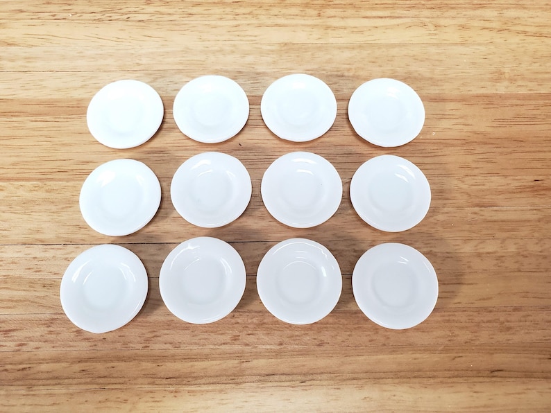 Pu&ograve; includere: Un set di dodici piccoli piattini circolari in ceramica bianca disposti in una griglia di tre file e quattro colonne. Ogni piattino presenta una finitura liscia e lucida con un centro leggermente incavato e un bordo rialzato uniforme.
