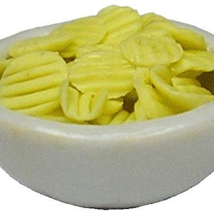 以下が含まれることがあります： 黄色いギザギザのポテトチップスが入った小さな白いセラミックボウル。チップスは薄い黄色で、作りたてのように見えます。ボウルは丸く、シンプルでクリーンなデザインです。ミニチュアの食品アイテム。