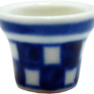 May include: A small ceramic cup with a white interior and a blue and white exterior design. The cup has a blue band near the top and a checkered pattern of blue squares. The cup has a flared shape.