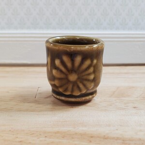 May include: A small, brown ceramic cup with a floral design. The cup is cylindrical with a slightly flared rim and sits on a wooden surface. The floral pattern is embossed around the cup's exterior.