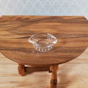 May include: A miniature clear glass bowl with a scalloped rim sits on a small, round wooden table. The table has a rich, warm wood grain finish and is supported by four turned legs. The background features a light-colored wall with a subtle floral pattern.