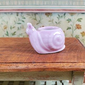May include: A small, glossy, light pink ceramic figurine shaped like a snail. The snail has a smooth, rounded shell with a visible spiral pattern and a small, raised head. The figurine features a hollow opening on the top of the shell, creating a miniature container or planter design.