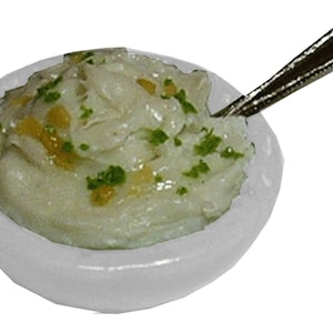 May include: A small white bowl filled with a light green, creamy food, garnished with green herbs and yellow specks. A silver spoon rests in the bowl. The food appears to be a dip or side dish.