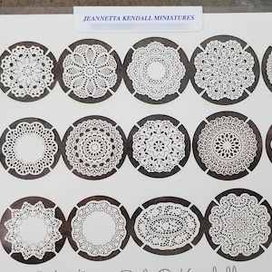 May include: A sheet of paper featuring various white lace doily designs in oval and circular shapes, each set within a dark gray circle. The text "JEANNETTA KENDALL MINIATURES" is printed at the top. These miniature doilies are ideal for craft projects.
