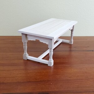 Op de afbeelding: Een kleine, witte houten tafel met een rechthoekig blad en vier sierlijke poten. De tafel is wit geschilderd en lijkt een miniatuur- of poppenhuismeubel te zijn. De tafel is ongeveer 30 cm lang.