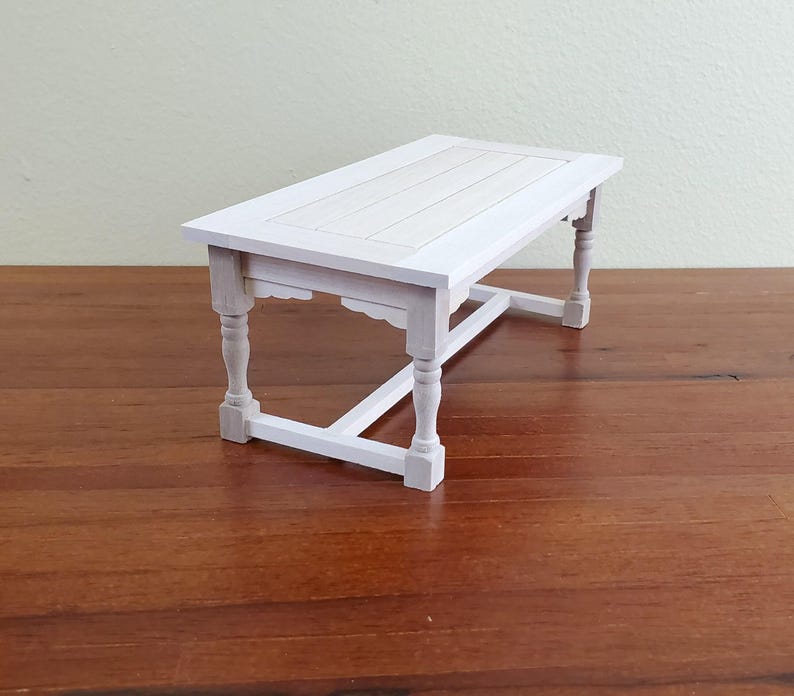 Op de afbeelding: Een kleine, witte houten tafel met een rechthoekig blad en vier sierlijke poten. De tafel is wit geschilderd en lijkt een miniatuur- of poppenhuismeubel te zijn. De tafel is ongeveer 30 cm lang.