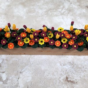 May include: A wooden planter box filled with artificial flowers in shades of orange, yellow, and burgundy, with green leaves. The rectangular planter is made of light-coloured wood. The arrangement is a vibrant display.