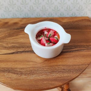 May include: A miniature white ceramic bowl filled with a red soup, garnished with white and green ingredients. The bowl has two handles and sits on a small, round wooden table. The table has a dark wood grain.