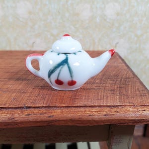 May include: A small, white ceramic teapot with a cherry design. The teapot has a red handle, spout, and lid accent. The cherry design features two red cherries and green leaves. The teapot is displayed on a wooden surface.