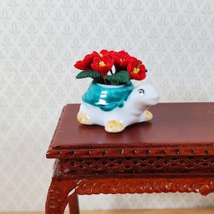 May include: A miniature ceramic turtle planter, white with a teal shell, holds vibrant red flowers. The turtle has yellow feet and is placed on a dark wooden table. The background features a light patterned wallpaper.