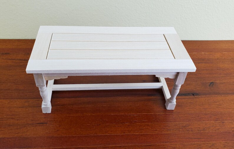 Op de afbeelding: Een kleine, rechthoekige witte houten tafel met een lattenbodem en sierlijke poten. De tafel is ongeveer 46 cm lang, 30 cm breed en 25 cm hoog. Hij staat op een donkerbruin houten oppervlak.