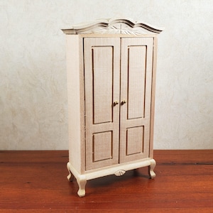 May include: A miniature wooden armoire with two doors, featuring a decorative top and brass-colored knobs. The armoire is painted in a light beige color and stands on four curved legs. The overall height is approximately 6 inches.