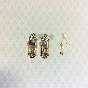 Könnte beinhalten: Miniatur-Türbeschlagsatz in Gold, bestehend aus zwei verzierten Türgriffen mit kristallartigen Mittelakzenten und zwei kleinen Schlüsseln. Die Türgriffe haben ein detailliertes, vintage Design. Die Schlüssel sind schlicht und elegant.