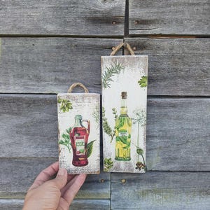 Könnte beinhalten: Zwei rechteckige Holztafeln mit Seilgriffen. Eine zeigt eine rote Flasche mit dem Wort "ESSIC" und Kräutern. Die andere hat eine gelbe Flasche mit der Aufschrift "BASIL OIL" mit einem Korken und Kräutern. Beide vor einem verwitterten Holzgrund.