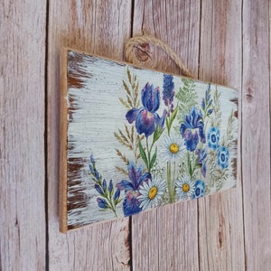 May include: A wooden sign with a distressed white background featuring a floral design with blue and purple irises and white daisies.