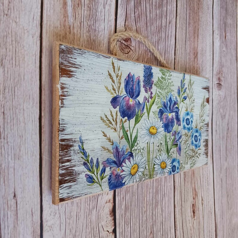 May include: A wooden sign with a distressed white background featuring a floral design with blue and purple irises and white daisies.