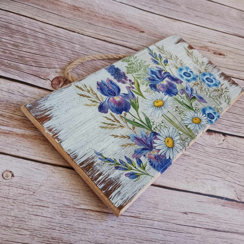 May include: A wooden sign with a white background and a floral design featuring blue irises and white daisies. The sign has a distressed, rustic look and is painted with a white wash. It has a rope hanger at the top.