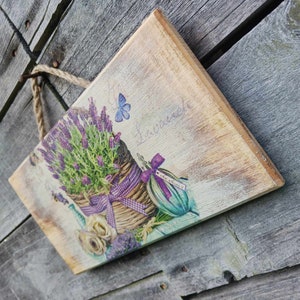 May include: A wooden sign with a lavender theme. The sign features a basket of lavender flowers, a butterfly, and a bottle of lavender oil. The text on the sign reads "La Lavande".
