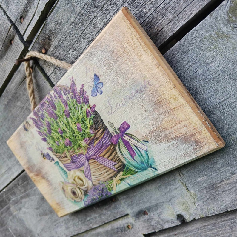 May include: A wooden sign with a lavender theme. The sign features a basket of lavender flowers, a butterfly, and a bottle of lavender oil. The text on the sign reads "La Lavande".