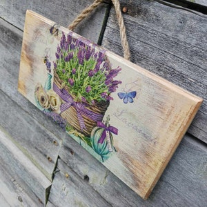 May include: A wooden sign with a lavender theme. The sign features a basket of lavender flowers, a butterfly, and the text "Fleur De Lavande".