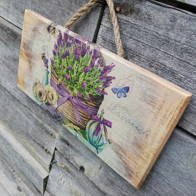 May include: A wooden sign with a lavender theme. The sign features a basket of lavender flowers, a butterfly, and the text "Fleur De Lavande".