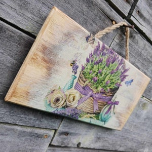 May include: A wooden sign with a vintage aesthetic featuring a basket of lavender flowers, white roses, and a blue watering can. The sign has a distressed, painted look and the text "Flower De..." is visible.