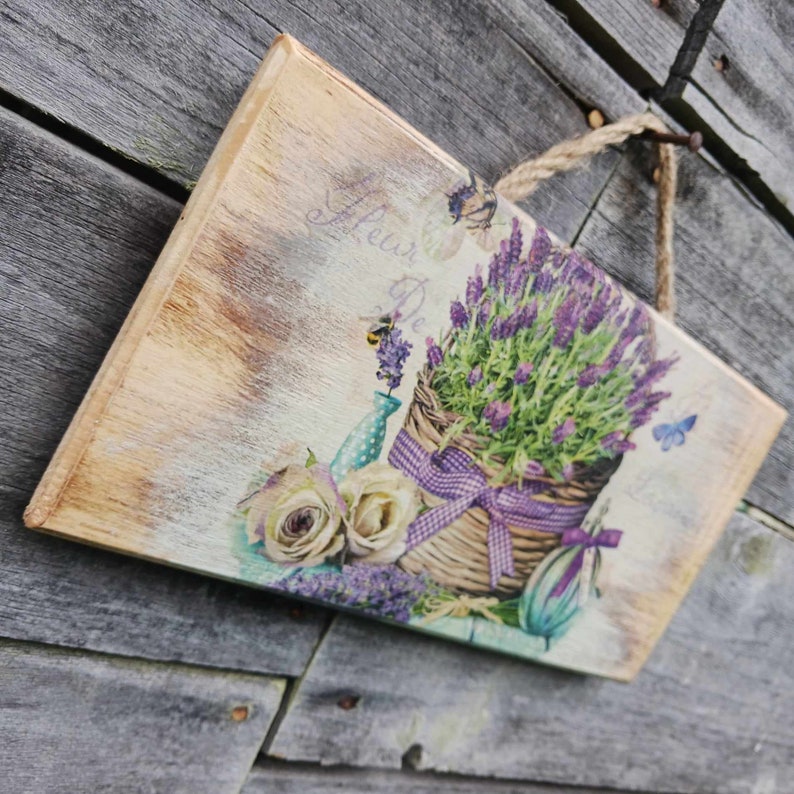 May include: A wooden sign with a vintage aesthetic featuring a basket of lavender flowers, white roses, and a blue watering can. The sign has a distressed, painted look and the text "Flower De..." is visible.