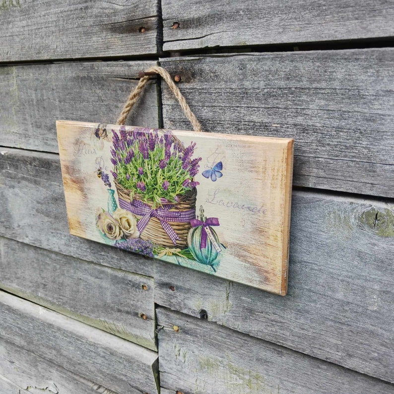 May include: A rustic wooden sign with a lavender floral design. The sign features a basket of lavender flowers, a butterfly, and the text "Fleur de Lavande".