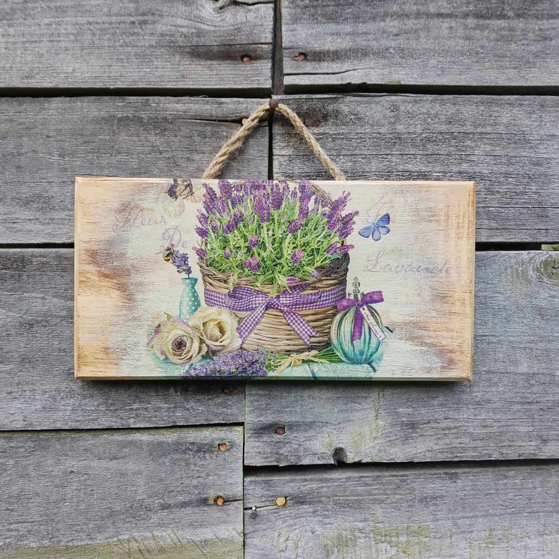 May include: A wooden sign with a distressed white background features a purple lavender plant in a wicker basket with a purple ribbon. The sign also includes a small bottle with a purple stopper and a butterfly. The text "Fleur De Lavande" is written on the sign.