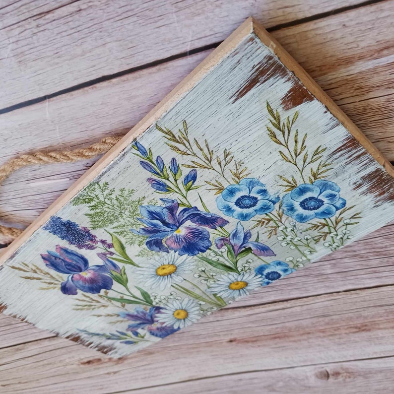 May include: A rustic wooden sign with a distressed white paint finish. The sign features a floral design with blue and purple irises, white daisies, and blue anemones. The sign has a rope hanger.