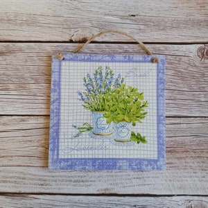 Puede incluir: Una impresión cuadrada enmarcada de color lavanda con fondo blanco. La impresión presenta una ilustración de acuarela de dos hierbas en maceta, lavanda y albahaca. El texto "Lavender" y "Basil" está escrito en cursiva en la impresión.