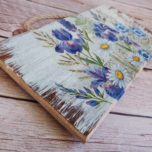 May include: A white wooden sign with a distressed finish features a floral design with blue and purple irises and white daisies. The sign has a rope hanger.