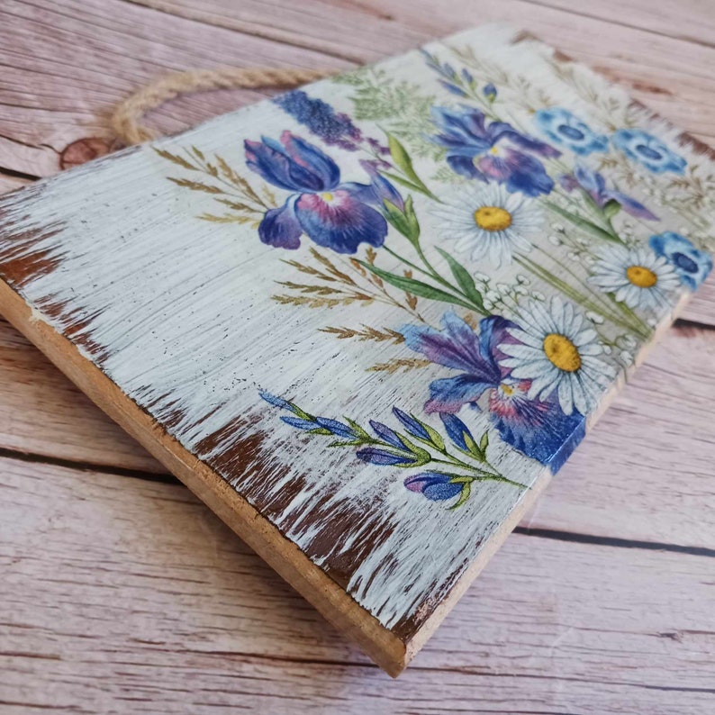 May include: A white wooden sign with a distressed finish features a floral design with blue and purple irises and white daisies. The sign has a rope hanger.