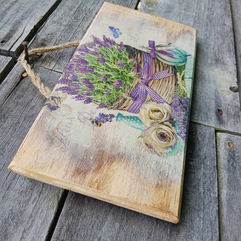 May include: A wooden sign featuring a lavender bouquet in a wicker basket, a butterfly, and the text "Fleur De". The sign has a distressed, rustic look and is ideal for farmhouse decor.