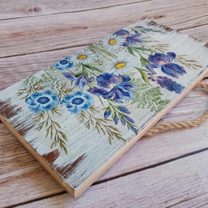 May include: A wooden sign with a distressed white paint finish features a floral design with blue, purple, and white flowers. The sign has a rope hanger.
