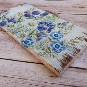 May include: A wooden sign with a distressed white paint finish features a floral design with blue and purple flowers. The sign has a rope hanger.
